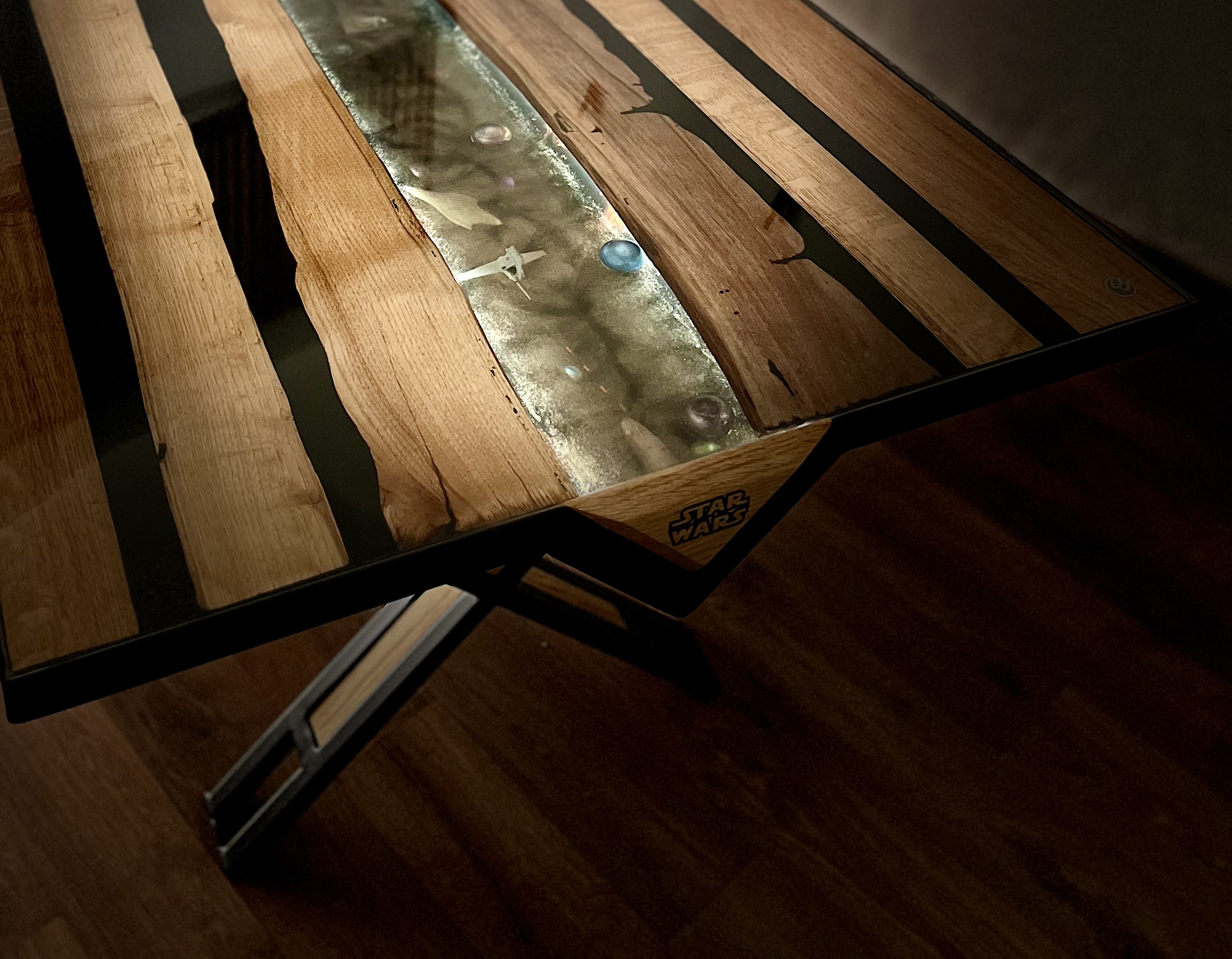 Table Starwars, pieds en métal recouvert de peinture mate, plateau en bois avec rivière époxy transparente. L'intérieur de la rivière est rétro éclairé par batterie qui met en évidence des vaisseaux.